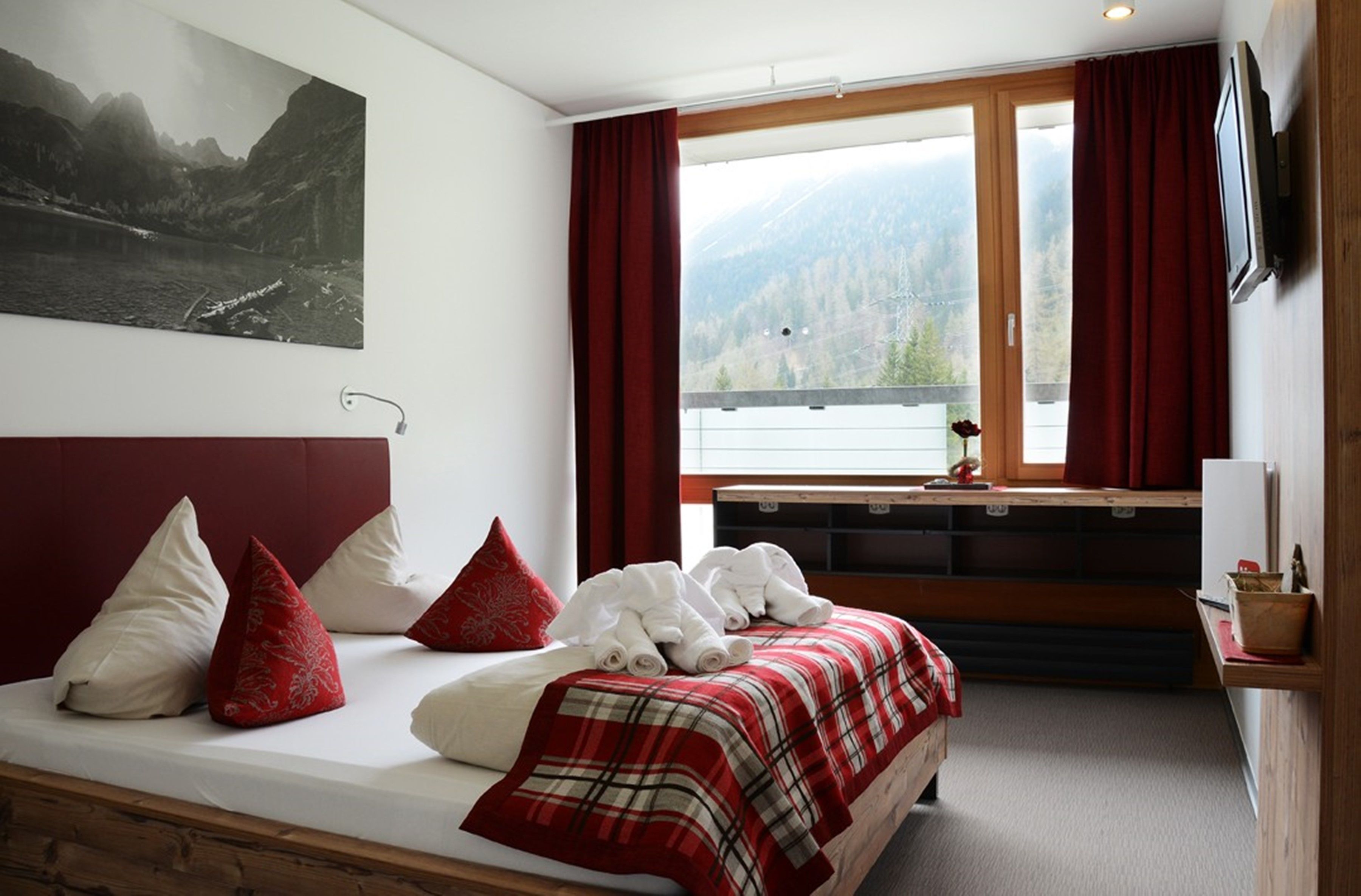 A hotel room with a bed, red curtains, a window with a mountain view, a mounted TV, and a painting on the wall.