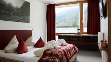 A hotel room with a bed, red curtains, a window with a mountain view, a mounted TV, and a painting on the wall.