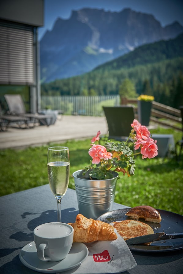 A table with a cup of coffee, a glass of white wine, a croissant, and a pastry on a plate, with a metal bucket of flowers and a scenic mountain view in the background.