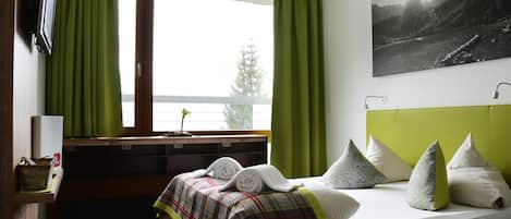 A bedroom with a bed, green curtains, a mounted TV, a painting, and a window with a view.