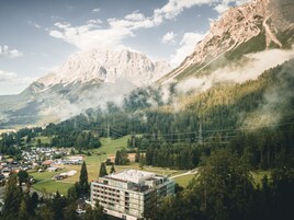 Un edificio moderno con techo plano y ventanas amplias, rodeado de exuberantes árboles verdes y montañas al fondo.