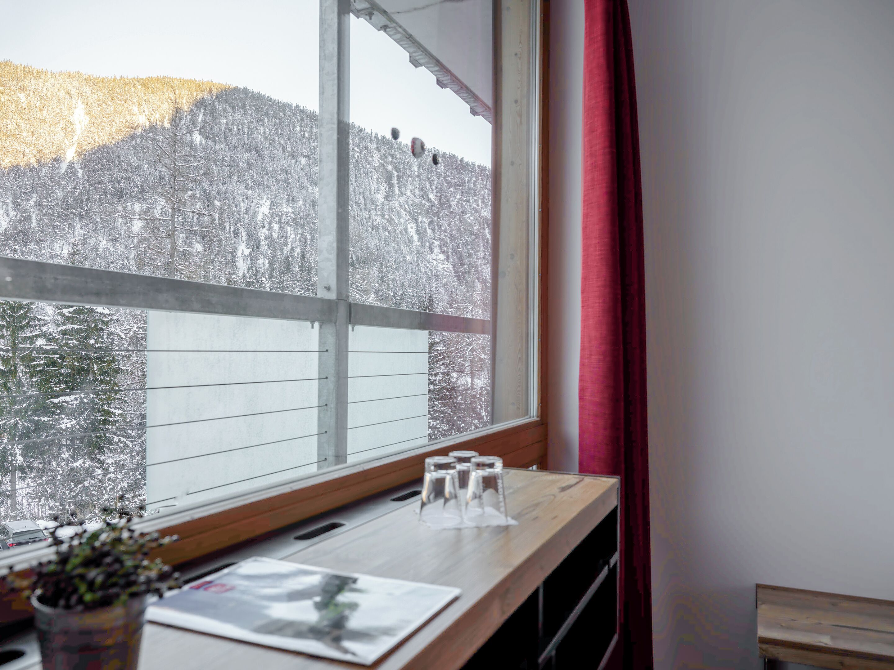 Una habitación con un ventanal grande que da a un bosque nevado. Hay una mesa de madera con un florero de vidrio y una revista encima.