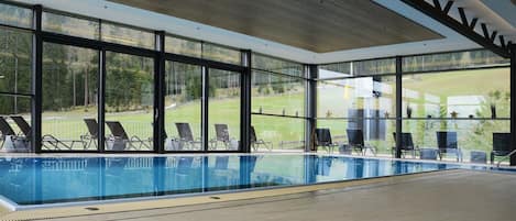 A large indoor swimming pool with clear glass walls, surrounded by lounge chairs, and a view of a green landscape.