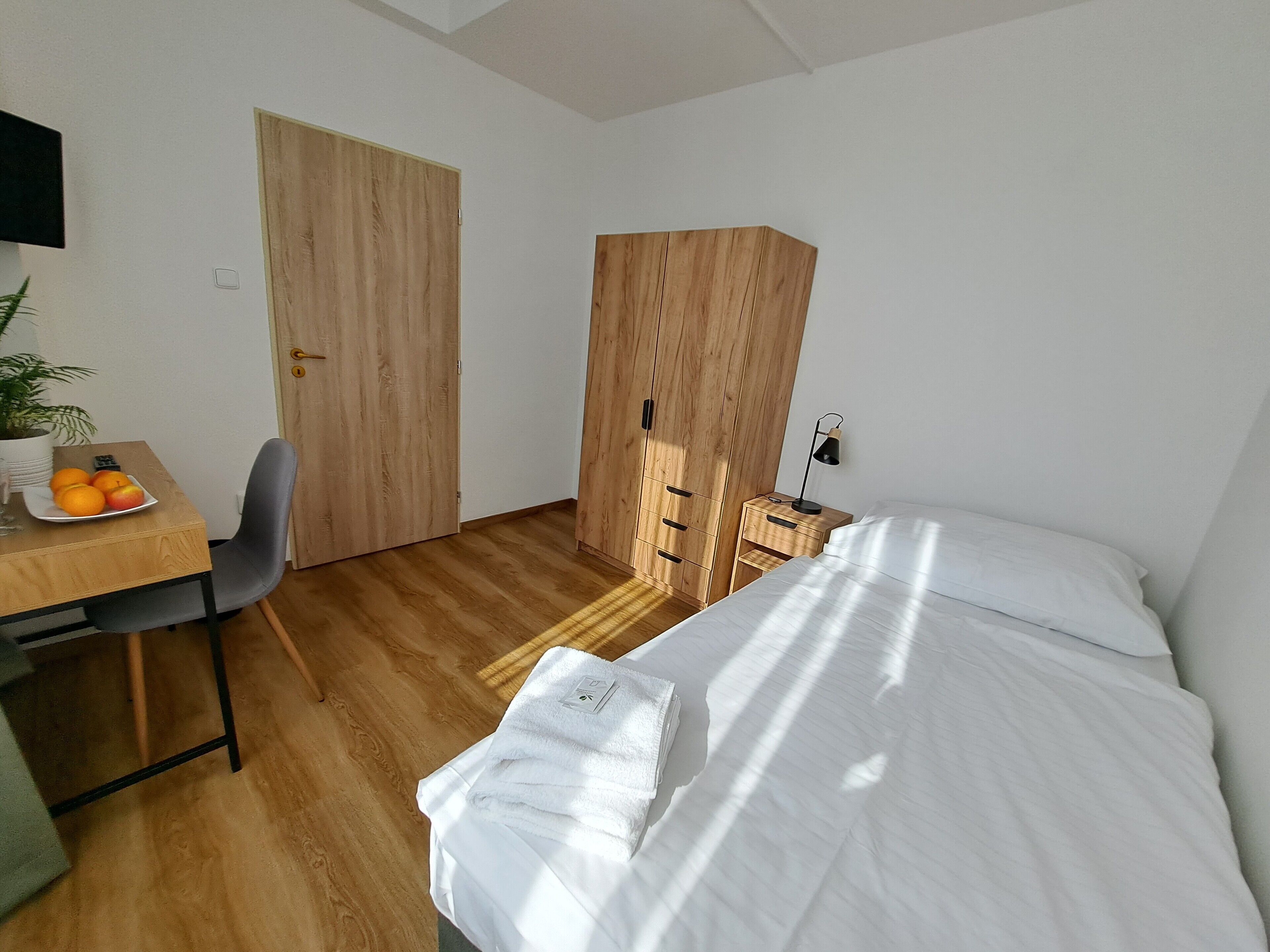 A small, modern bedroom with a wooden floor, a bed with white linens, a desk with a plate of fruit, a chair, a wardrobe, and a bedside table with a lamp.