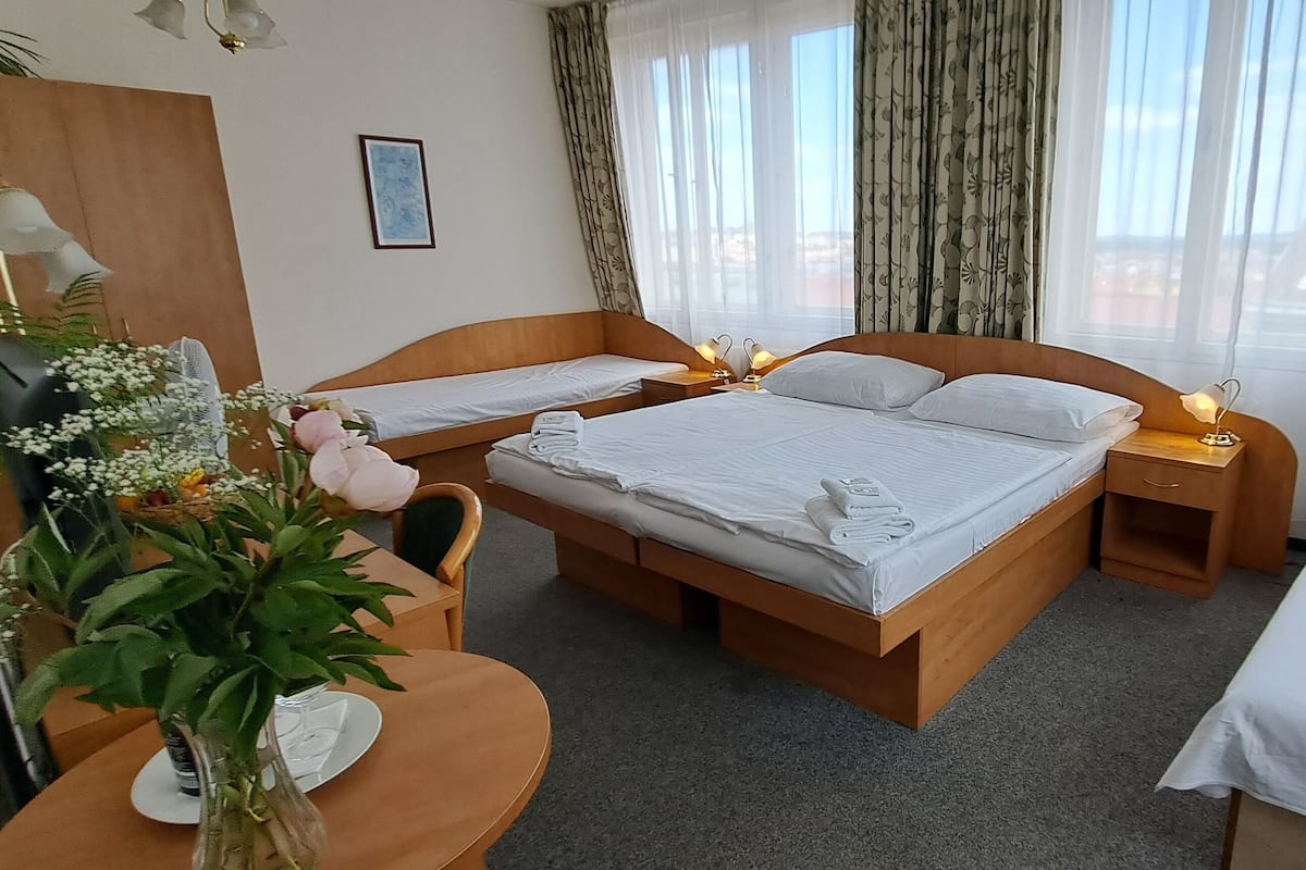 A hotel room with two single beds, a wooden desk with a chair, a small table with a vase of flowers, and a view of the city through large windows with sheer curtains.