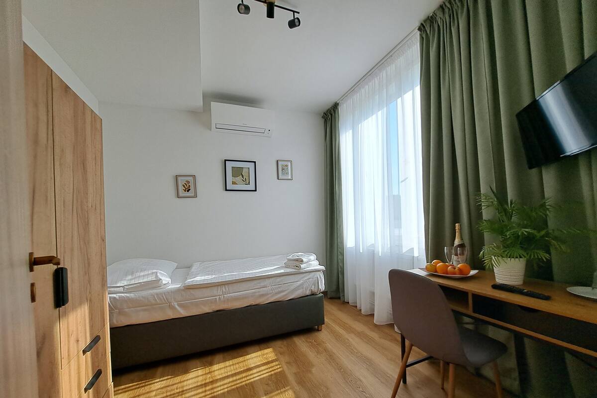 A modern hotel room with a bed, a desk with a chair, a television, and a window with curtains.