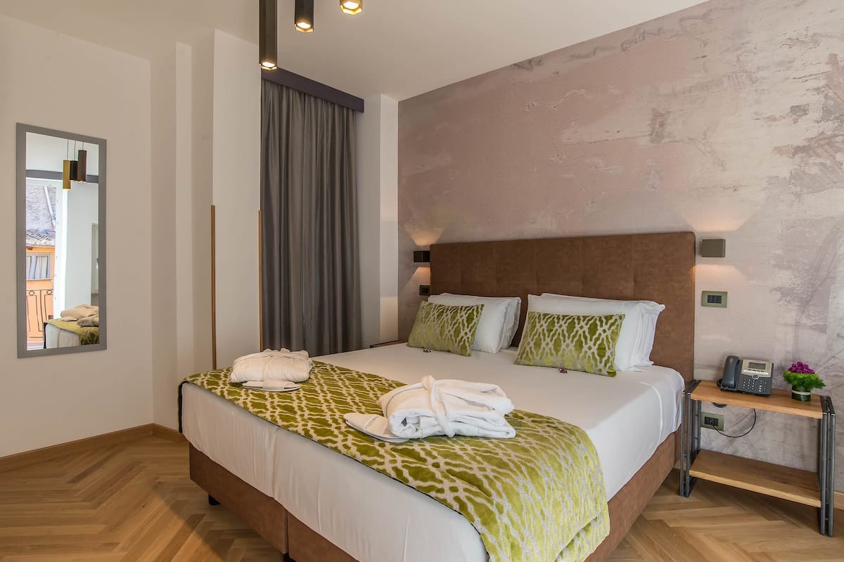 A neatly made bed with a patterned bedspread, a bedside table with a phone and a small plant, and a mirror on the wall.