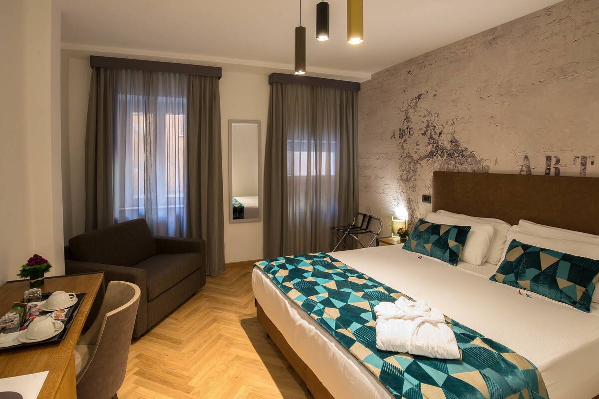 A hotel room with a large bed, a sofa, a dining table with breakfast items, and a view of the city through the window.