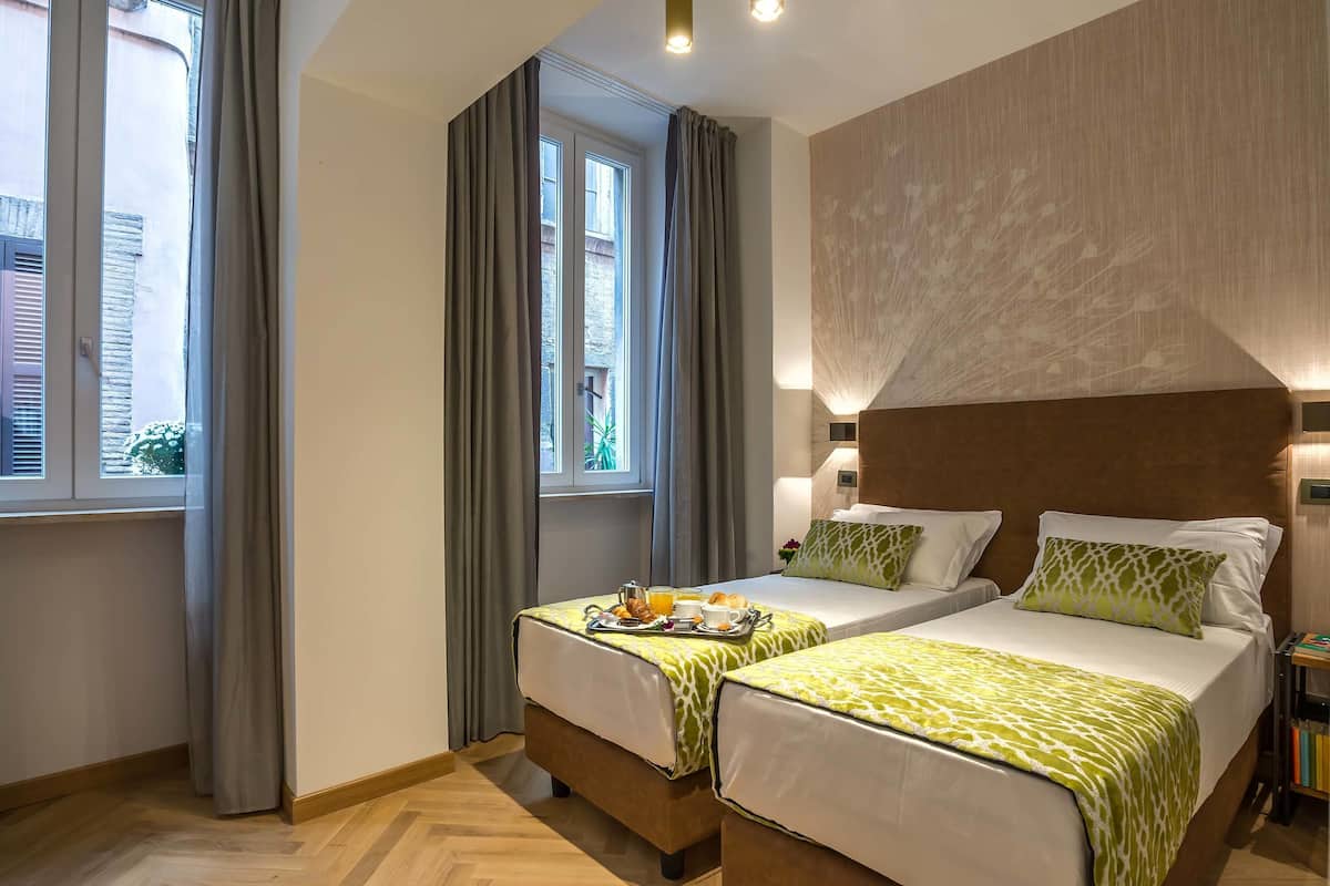 A hotel room with two beds, a large window with curtains, a wooden headboard, and a bedside table with a lamp.
