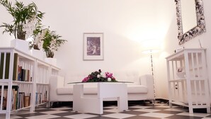 Una habitación con piso a cuadros, sofás blancos, una mesa de centro de vidrio, un cuadro enmarcado en la pared y estanterías blancas con libros y plantas.