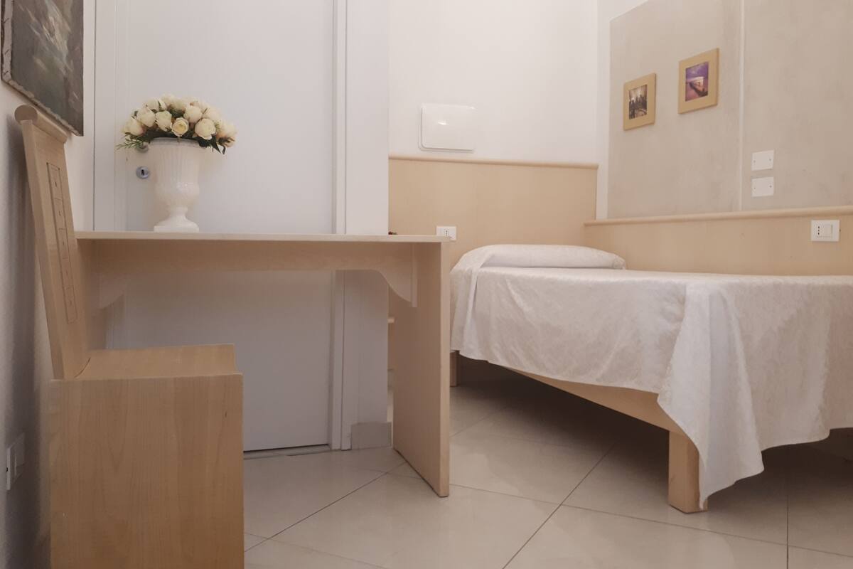 A small, neatly arranged room with a bed, a desk, and a wall-mounted shelf with a vase of flowers.