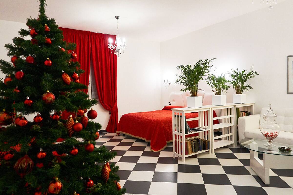 A modern bedroom with a checkered floor, a red bedspread, a Christmas tree, and a white console table with books.