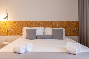 Una cama bien tendida con sábanas blancas y dos toallas enrolladas, un cabecero con textura de corcho y una mesita de noche con lámpara.