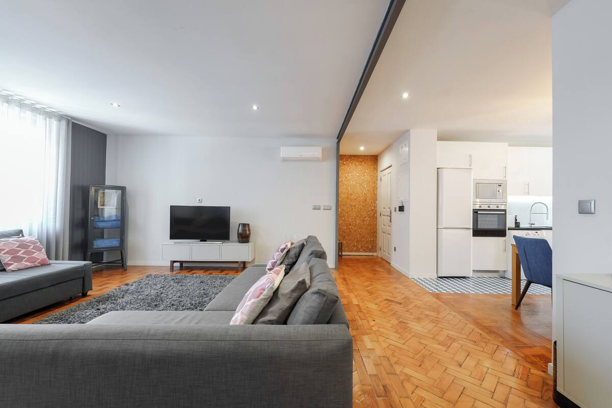 Una sala de estar moderna con un sofá gris, un televisor de pantalla plana y una cocina al fondo.