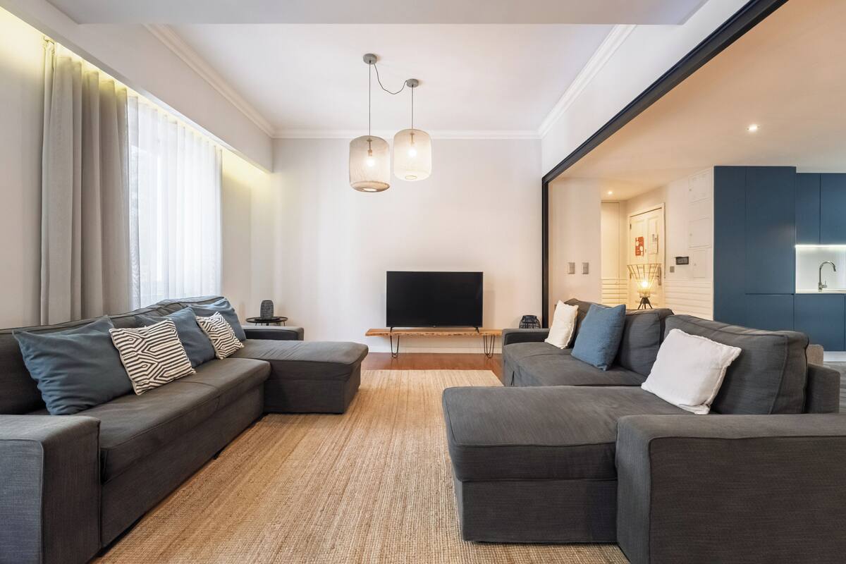 Una sala moderna con un sofá seccional, un televisor de pantalla plana y una cocina visible a través de una puerta abierta.