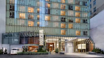 Un hotel moderno con fachada de vidrio, una entrada destacada y un restaurante visible llamado "BOQUERIA" en la planta baja.