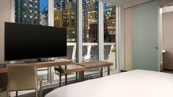 Habitación de hotel con una cama grande, un escritorio con televisor y vistas a edificios de la ciudad a través de amplias ventanas.