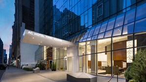 L’extérieur de l’hôtel AC Times Square se distingue par sa façade moderne en verre, son entrée imposante et un espace détente visible à travers les fenêtres.