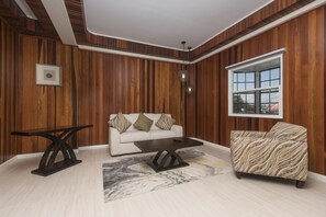A modern living room with a wooden paneled wall, a white sofa with patterned cushions, a patterned armchair, a dark coffee table, and a large window with a view. - King's Hotel & Residences (Georgetown)