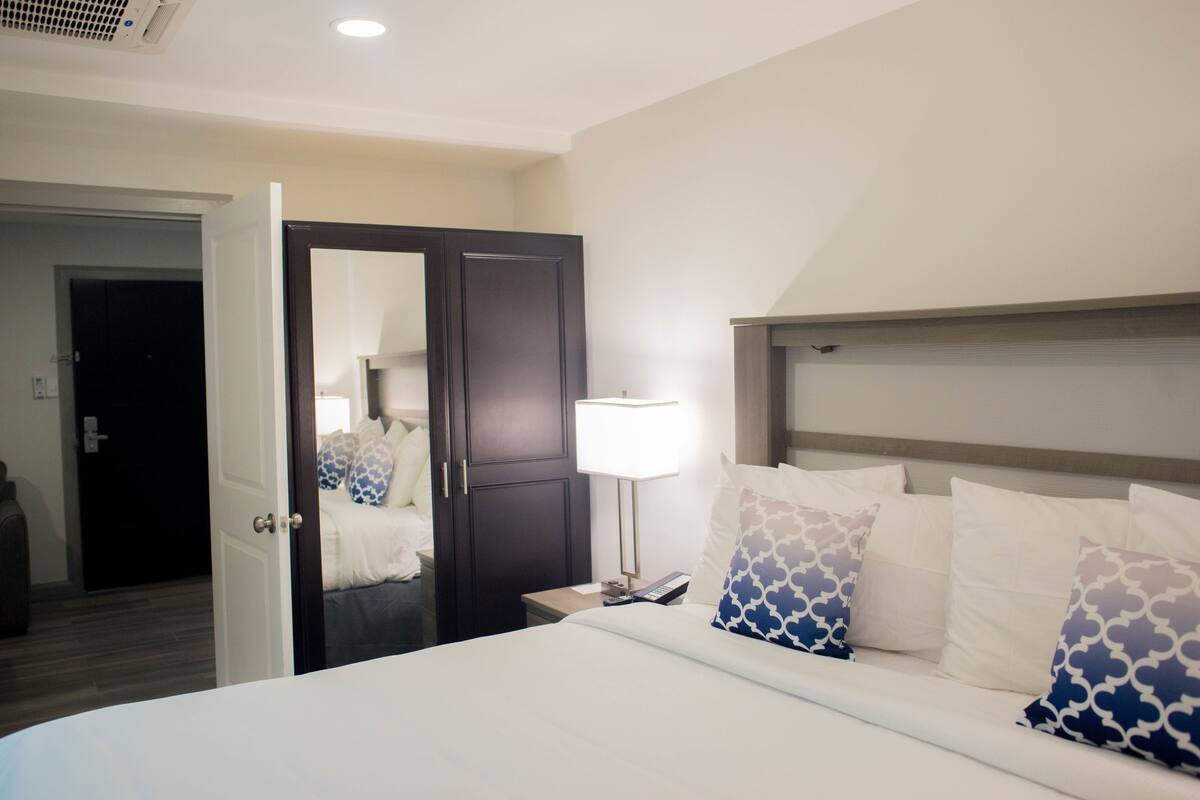 A hotel room with a bed, a nightstand, a lamp, and a door leading to another room.