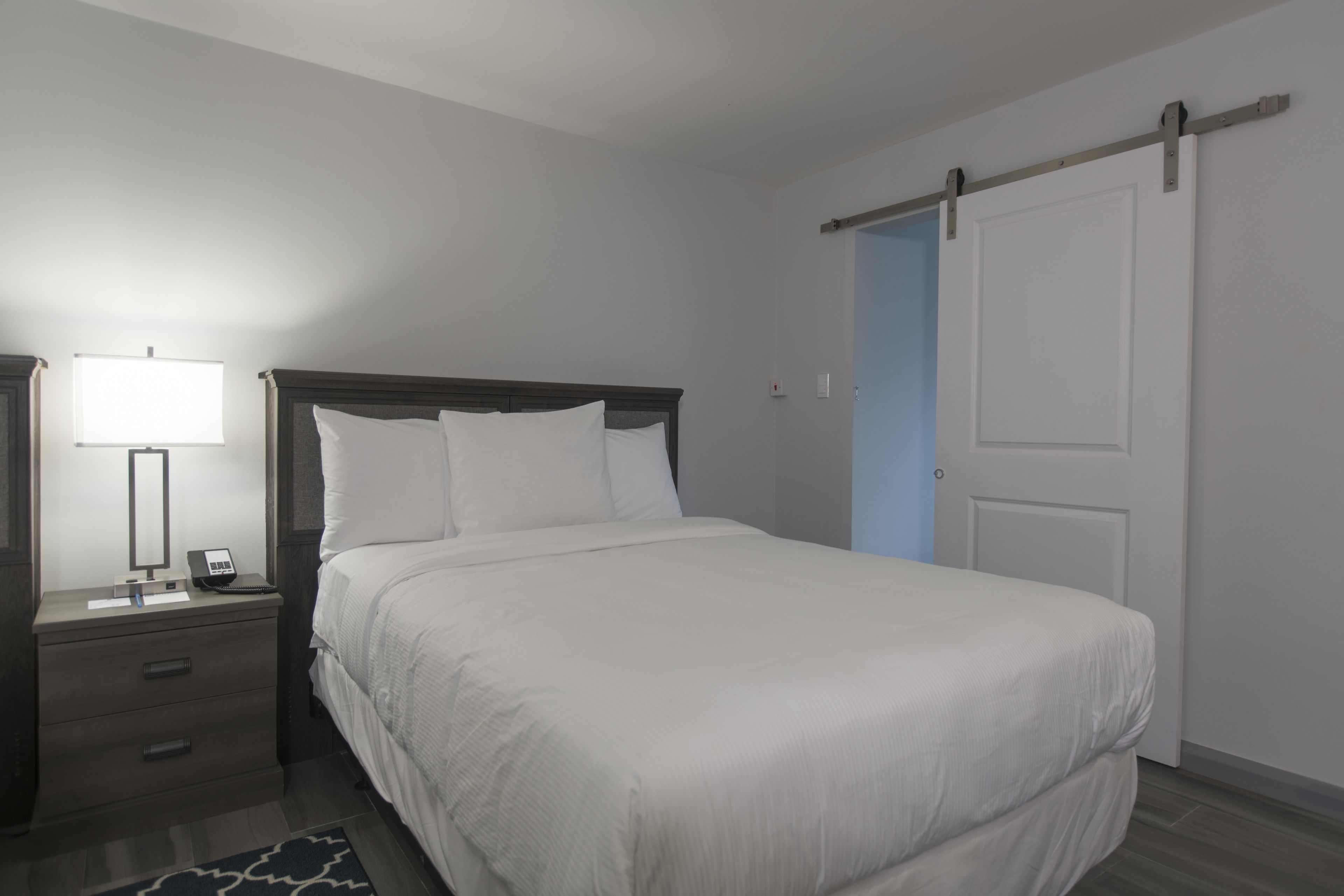 Een netjes opgemaakt bed met witte lakens, een nachtkastje met een lamp, en een deur met een schuifmechanisme.