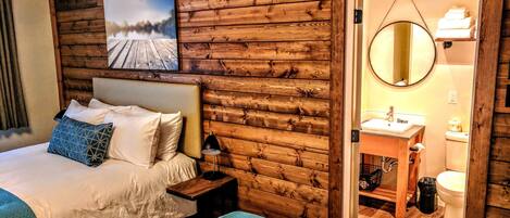 A wooden cabin-style bedroom with a bed, pillows, a nightstand, a bedside lamp, a wooden wall with a painting, and a bathroom visible through an open door.