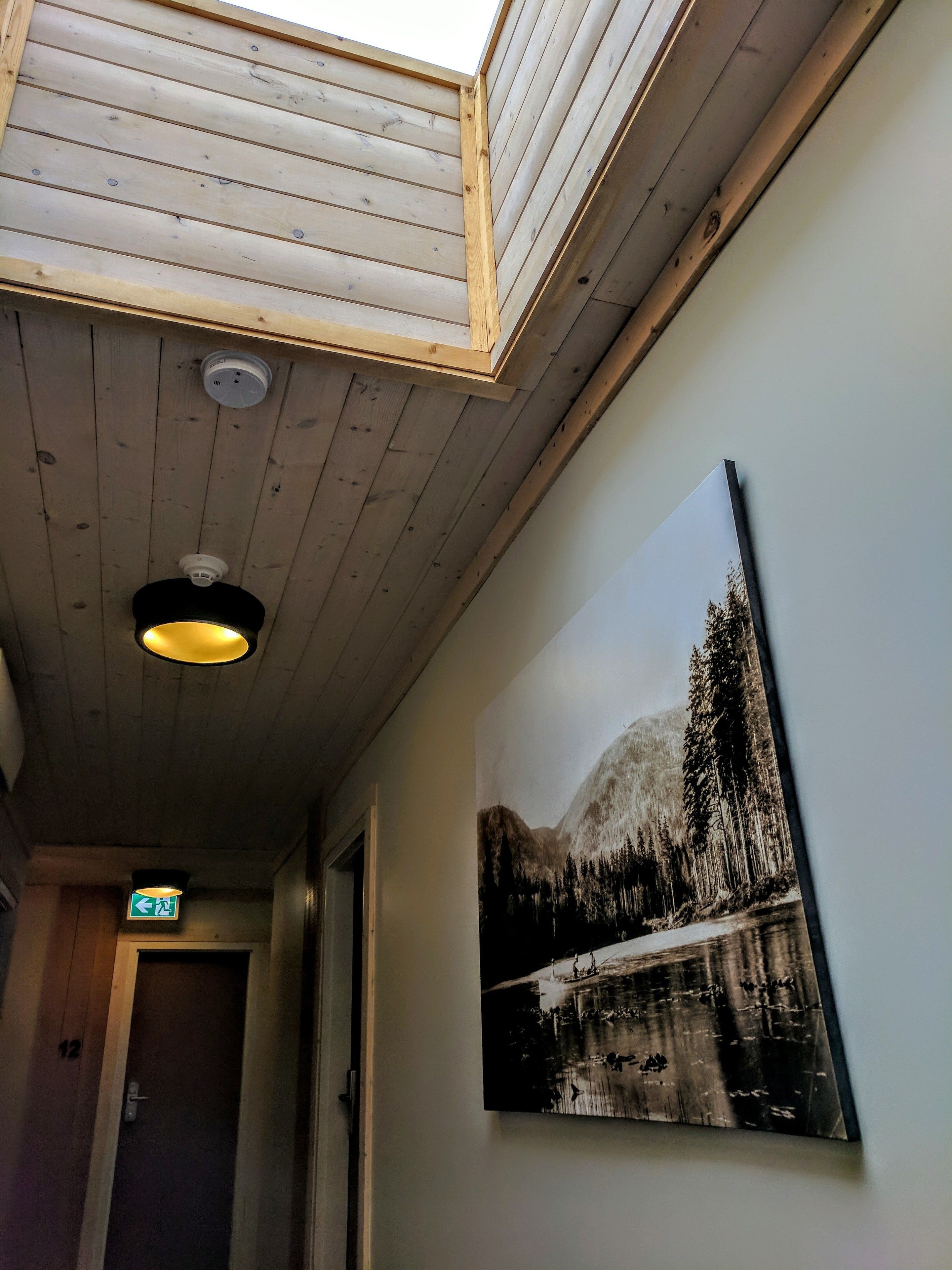 A wooden ceiling with skylight, a mounted picture of a mountain landscape, and a door with an exit sign.
