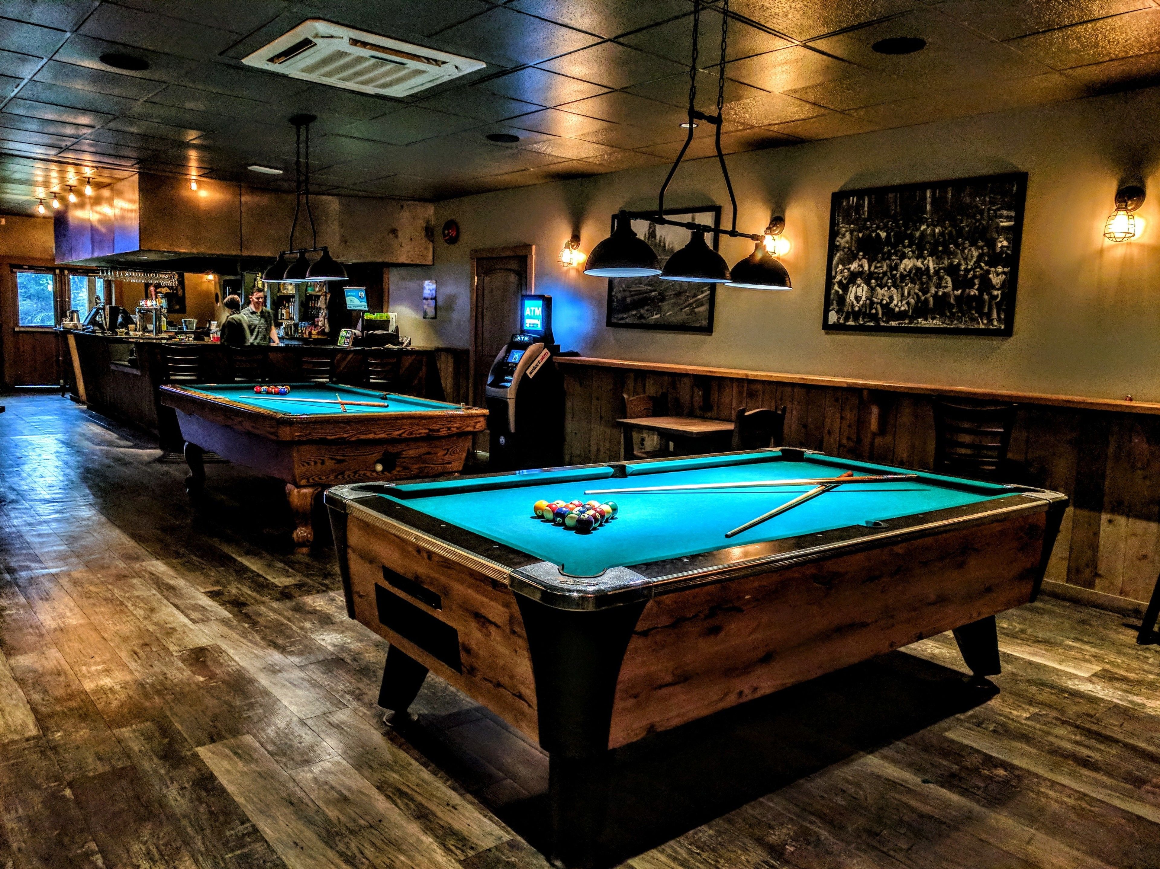 Een pooltafel met houten afwerking en een blauwe viltkleed, een bar met een toonbank en zitplaatsen, en een aan de muur bevestigde pinautomaat.