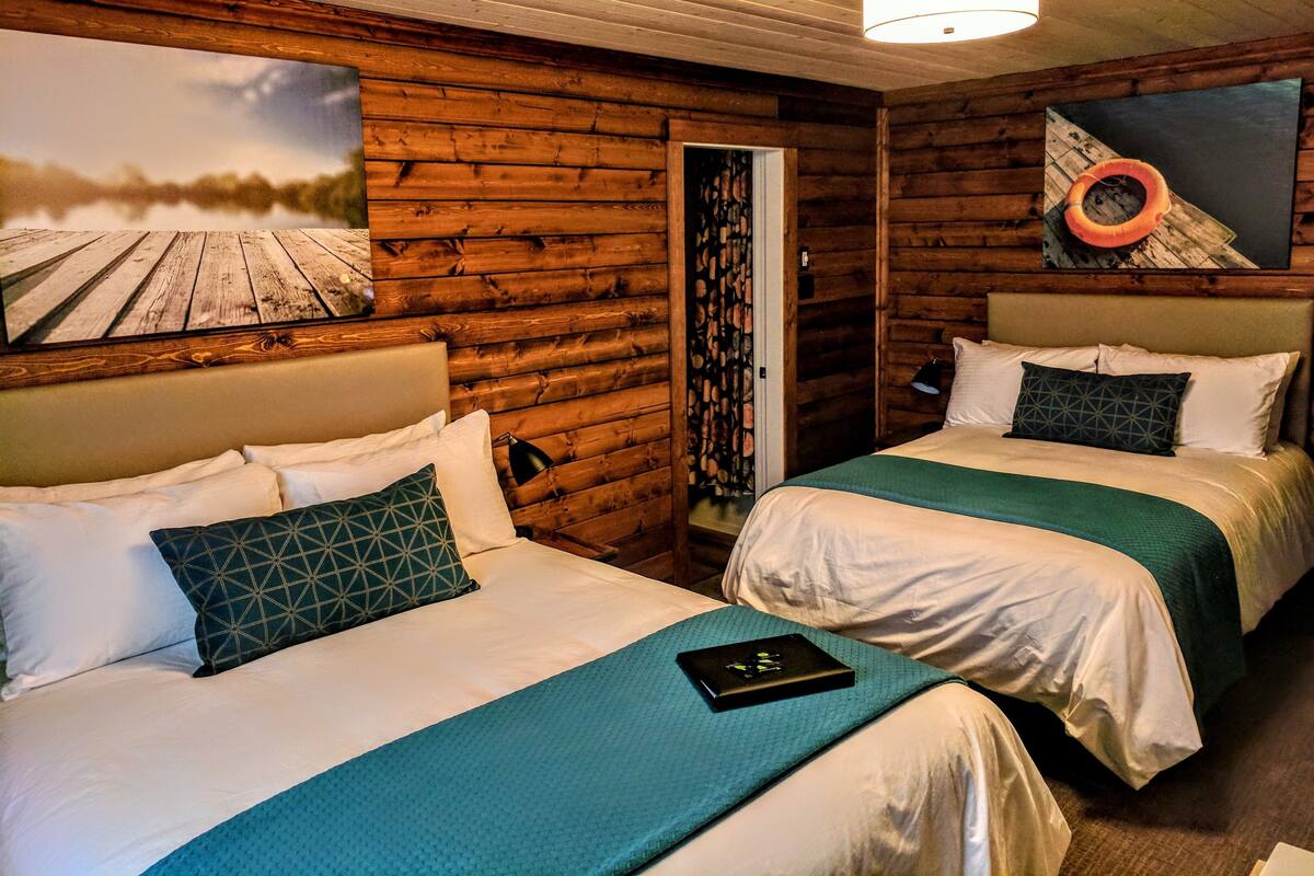 Habitación tipo cabaña con dos camas, paredes de madera, un cuadro de un barco y una lámpara en el techo.