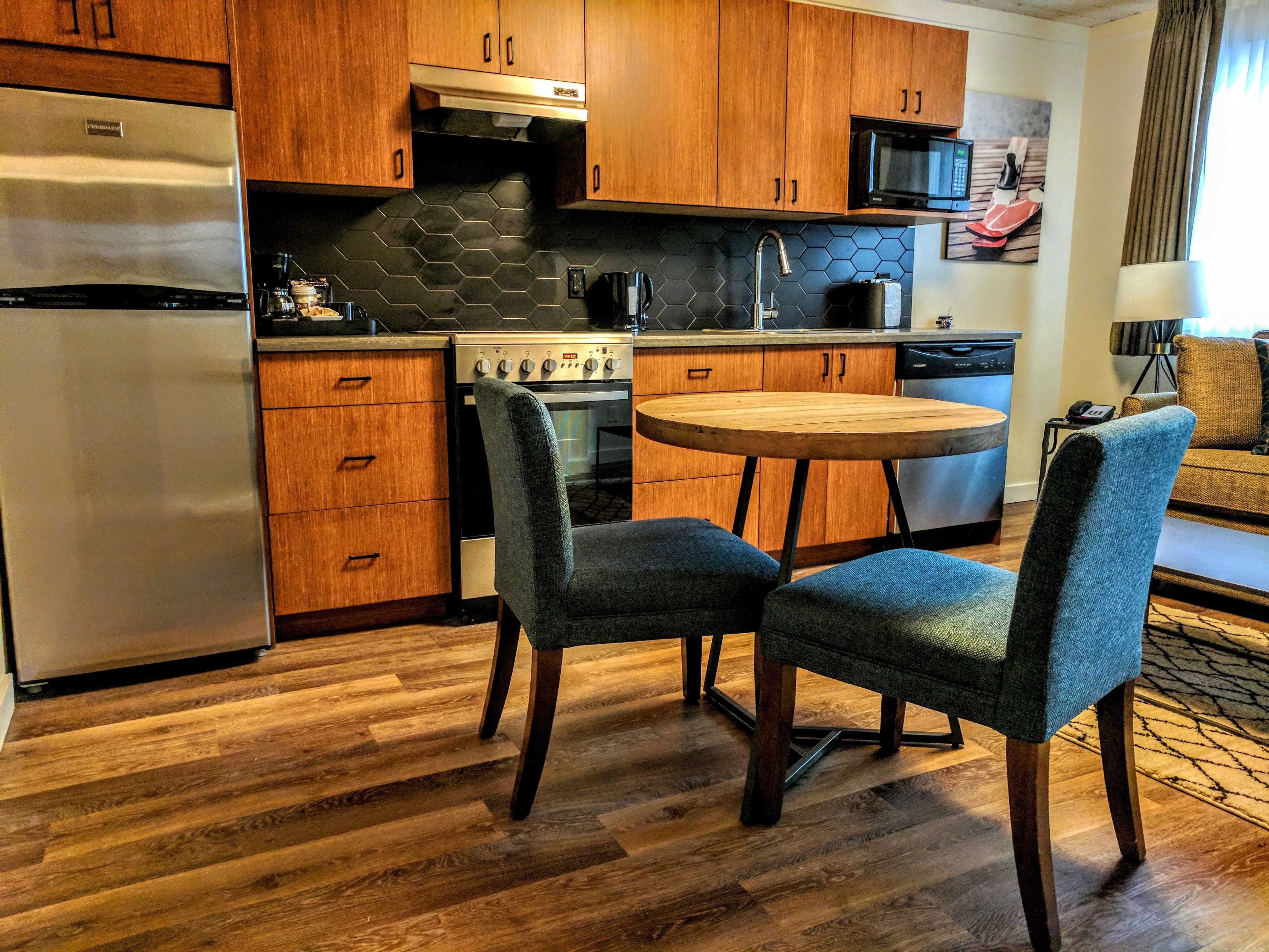 A compact kitchen with wooden cabinets, a stainless steel refrigerator, a microwave, and a stove. There is a round wooden table with two chairs.