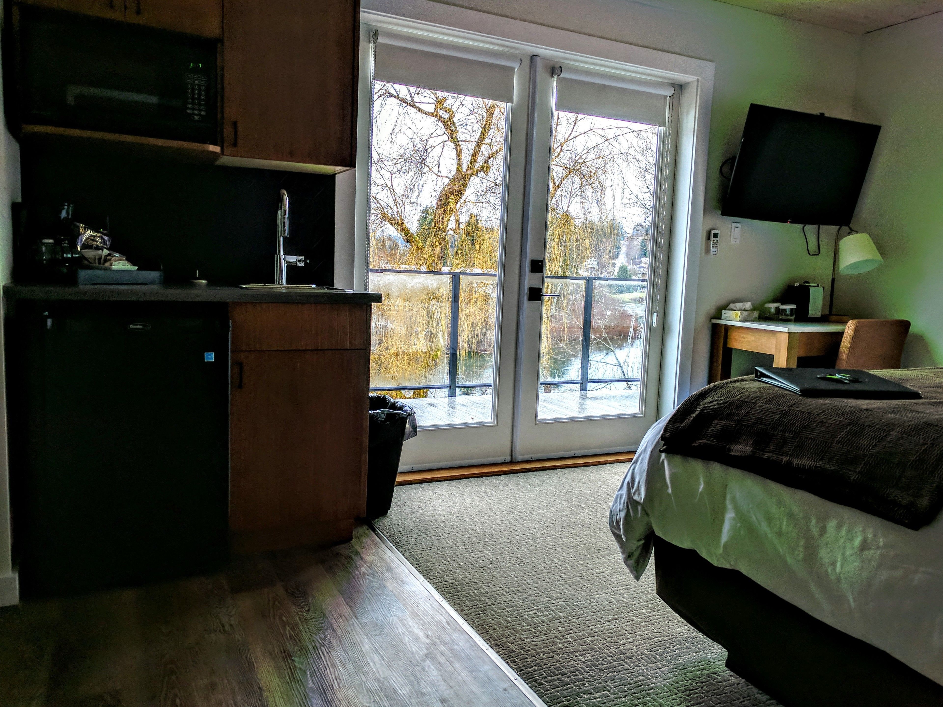 A room with a kitchenette, a bed, a TV, and a view of trees through a sliding glass door.