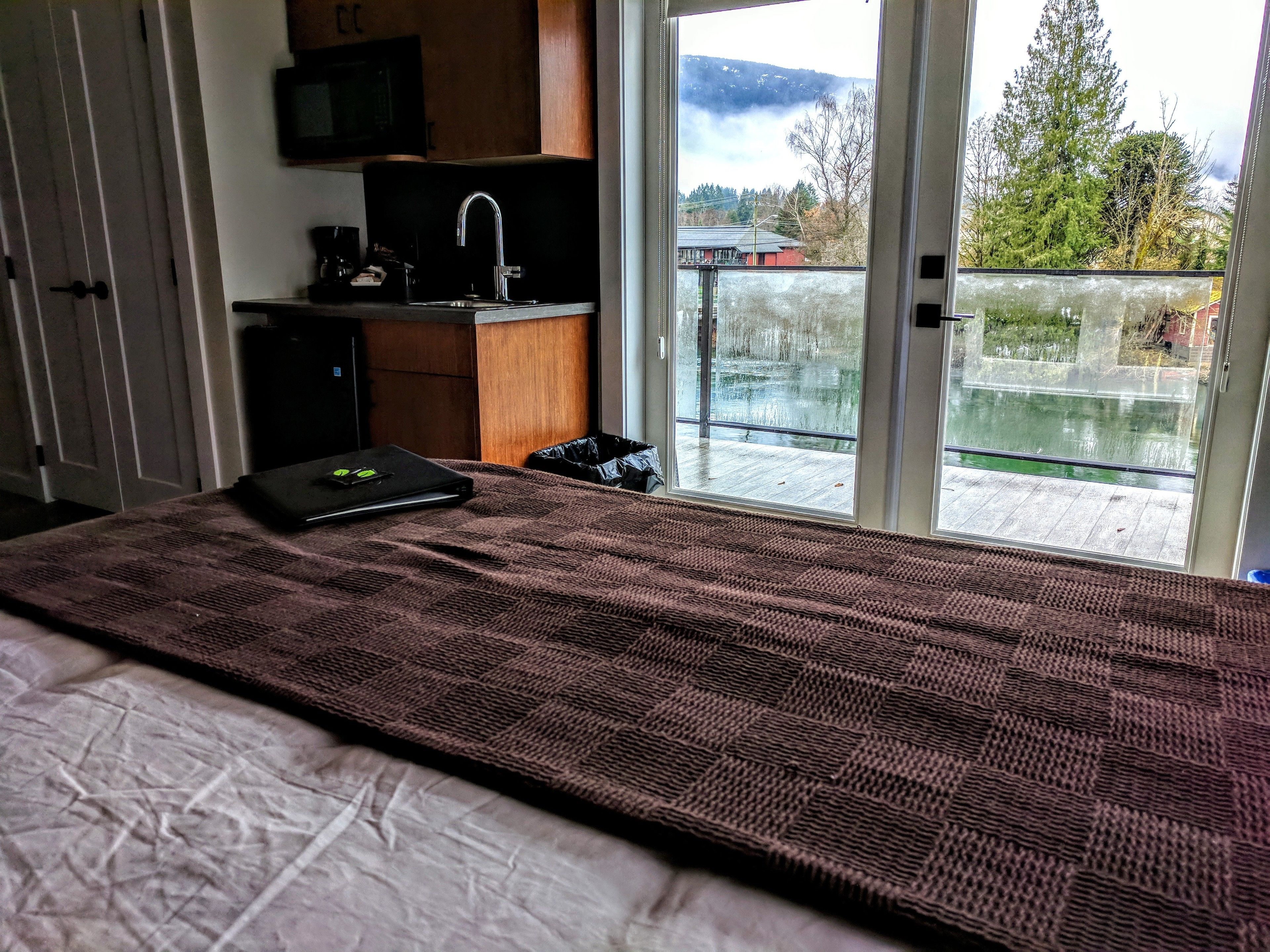 Een slaapkamer met een bed, een laptop, een wastafel en uitzicht op een balkon met een meer en bomen.