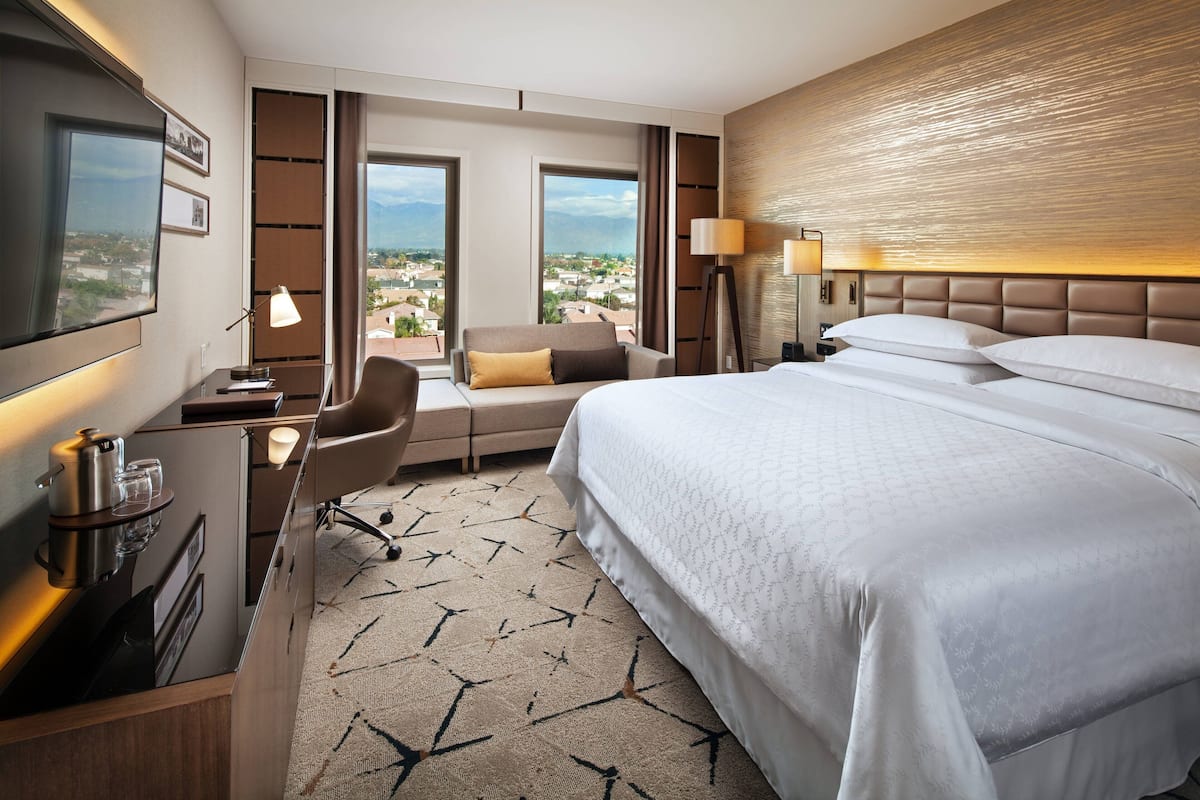 Hotelkamer met een groot bed, een bureau met lamp, een stoel en uitzicht op de stad door grote ramen.