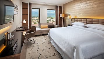 A hotel room with a large bed, a desk with a lamp, a chair, and a view of the city through large windows.