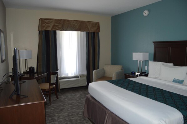 Habitación de hotel con cama, escritorio, silla, lámpara de escritorio, mesita de noche con lámpara y un reloj de pared.
