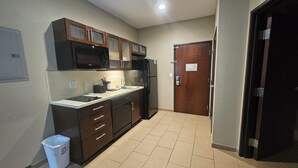 Cocina compacta con microondas, refrigerador y pileta. Hay un bote de basura y una puerta con un cartel.
