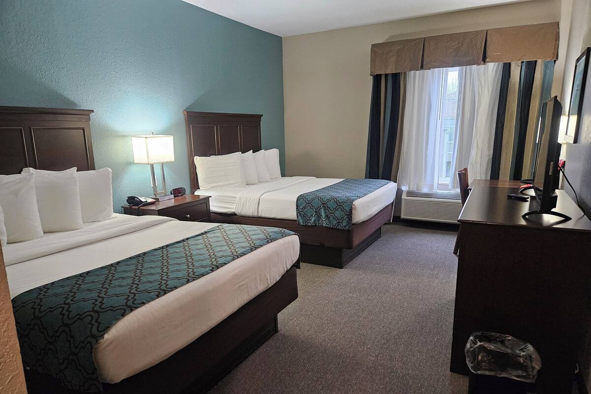Une chambre d’hôtel avec deux lits, un bureau, une chaise, une lampe et une fenêtre avec des rideaux.