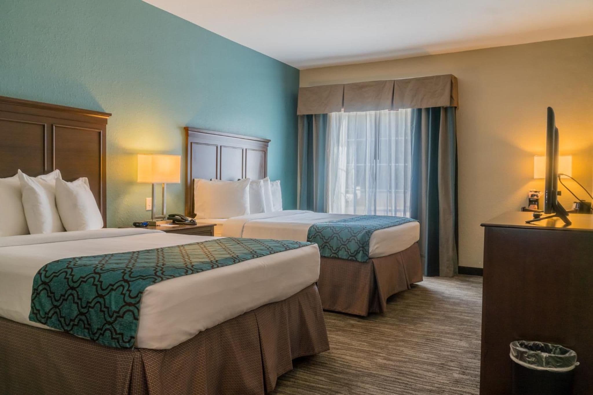 A hotel room with two beds, a desk, a lamp, and a window with curtains.