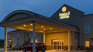 Entrada de un hotel con un área techada, un vehículo rojo estacionado al frente y un cartel que indica que es un Heritage Place Hotel & Suites.