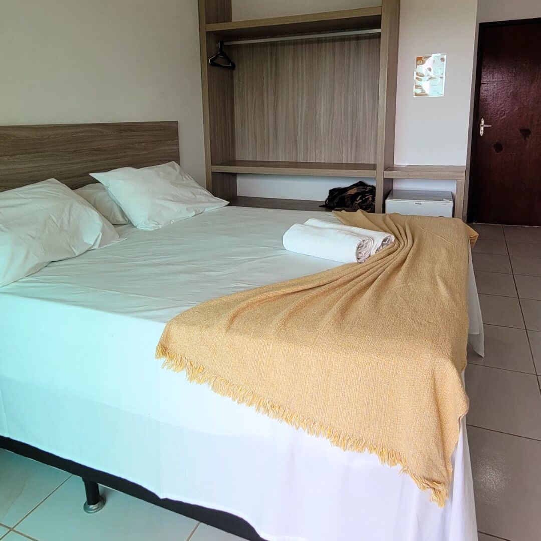 Ein ordentlich bezogenes Bett mit weißer Bettdecke und beiger Decke, ein Holz-Kopfteil, eine kleine Ablage mit Kleiderhaken und eine Tür im Hintergrund.