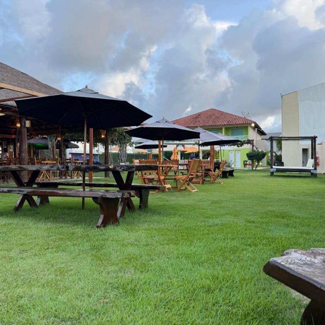 An outdoor dining area with wooden tables and umbrellas, surrounded by a grassy lawn and a building with a red roof.