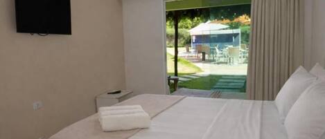 A hotel room with a bed, a TV, a window with curtains, and a view of an outdoor area.
