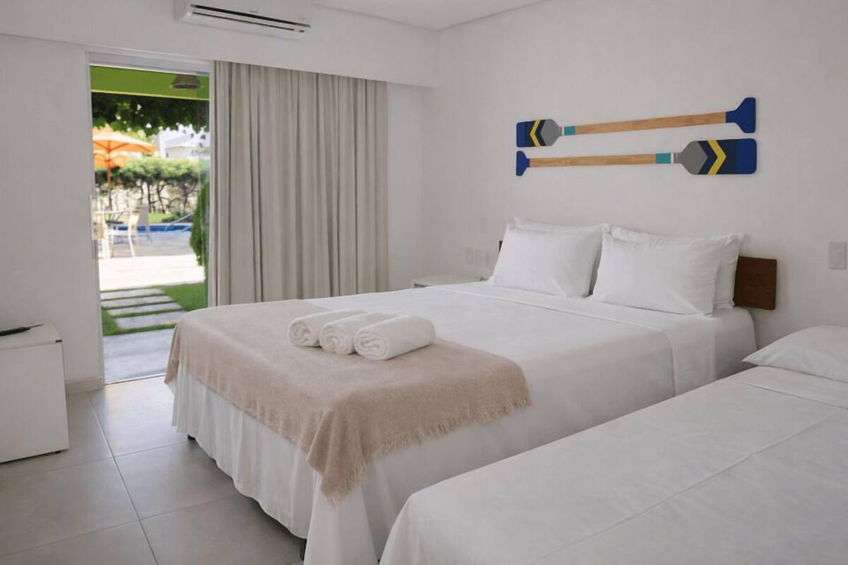 Une chambre d’hôtel comprenant un lit, une table de chevet, un climatiseur fixé au mur et donnant sur un espace extérieur avec une piscine et des parasols.