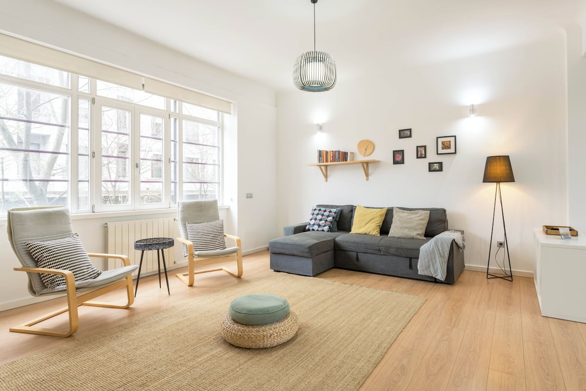 Ein modernes Wohnzimmer mit einem Ecksofa, zwei Sesseln, einem runden Hocker, einer Stehlampe und einer Pendelleuchte.