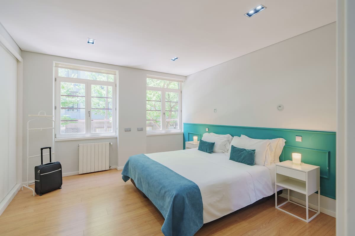 Ein modernes Schlafzimmer mit einem großen Bett, einem Nachttisch, einem Koffer und einem Fenster mit Blick auf Bäume.