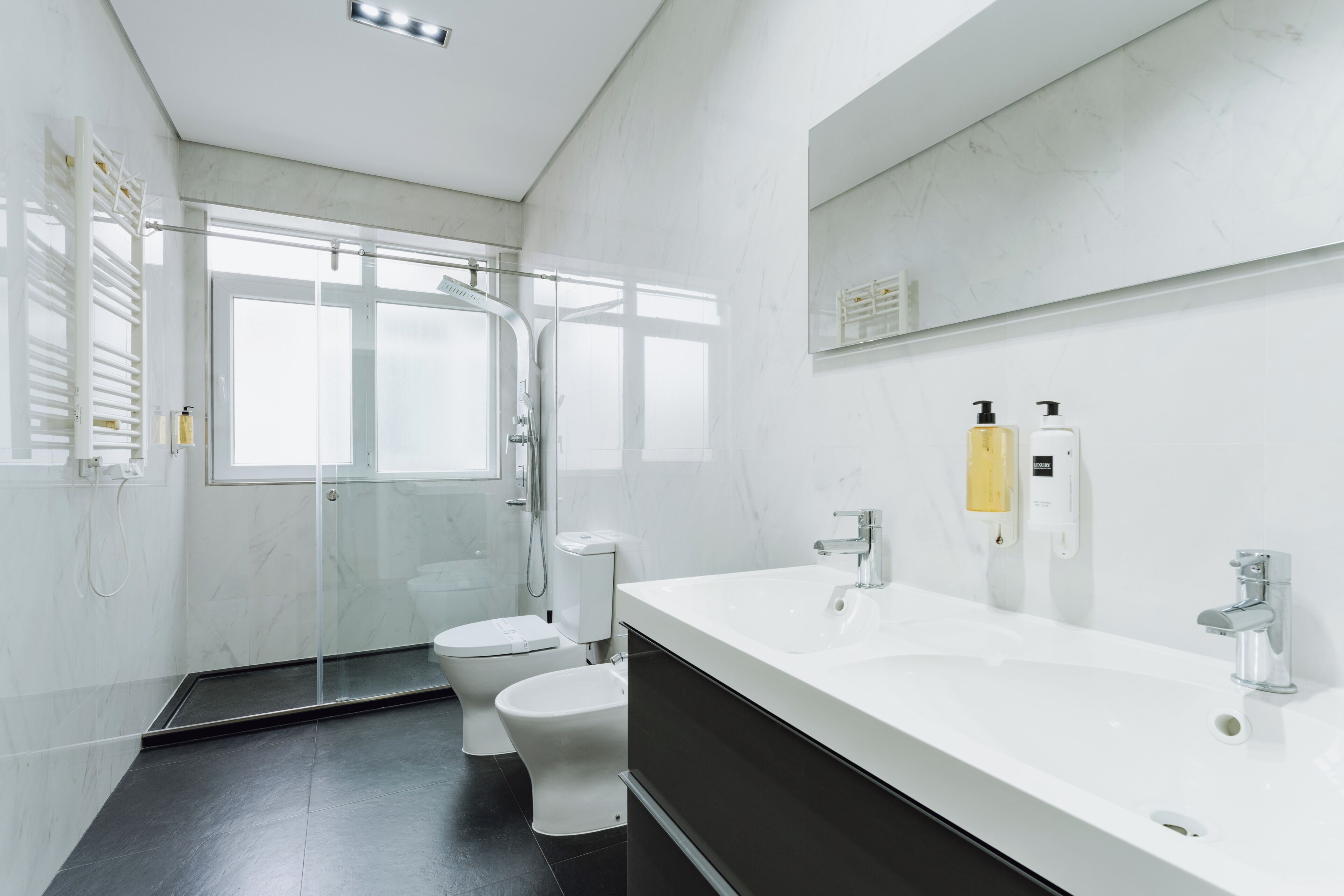 Um banheiro moderno com box de vidro, vaso sanitário branco, pia grande branca com espelho em cima e mueble escuro.