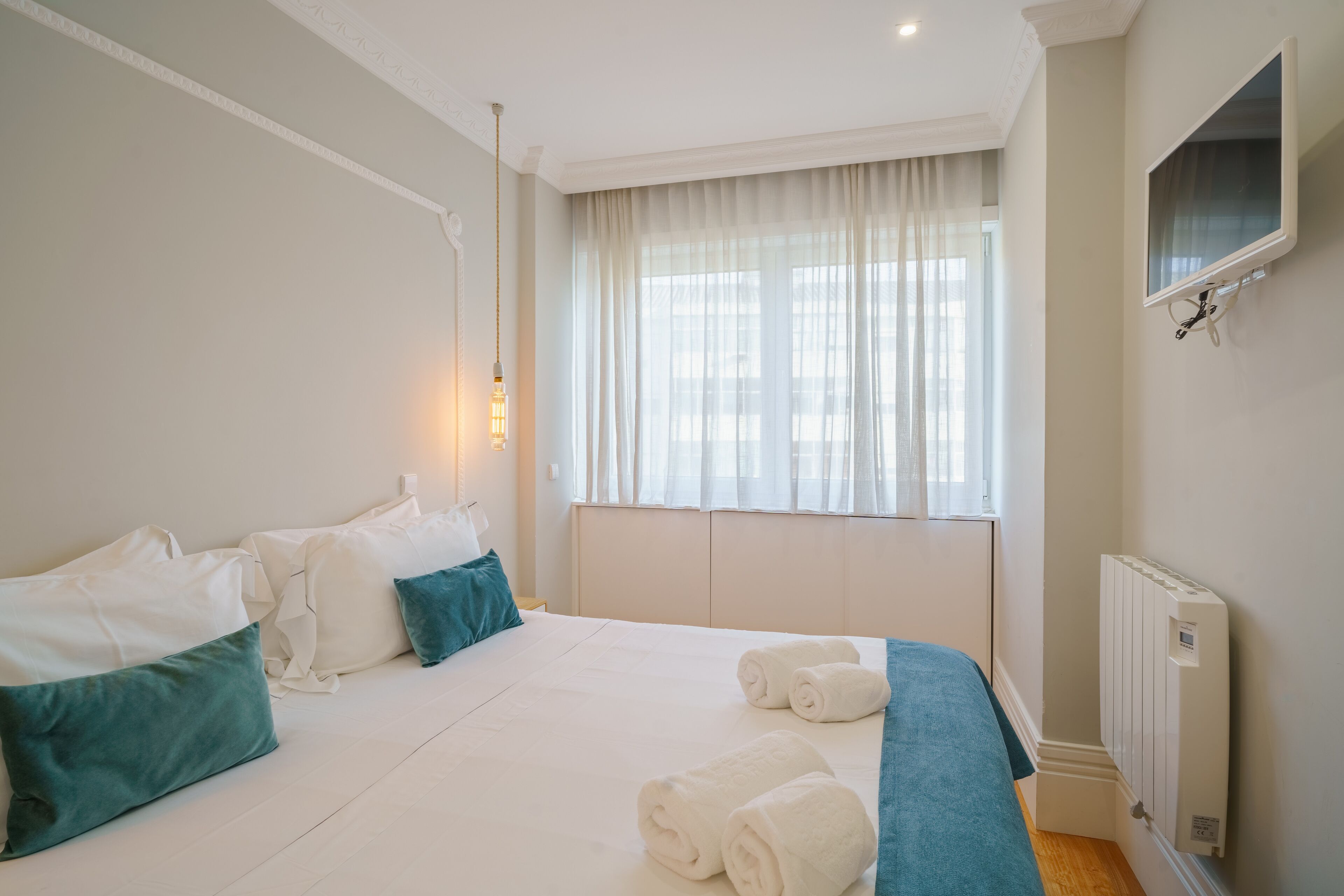 A hotel room with a large bed, white bedding, teal pillows, a window with sheer curtains, a wall-mounted TV, and a radiator.