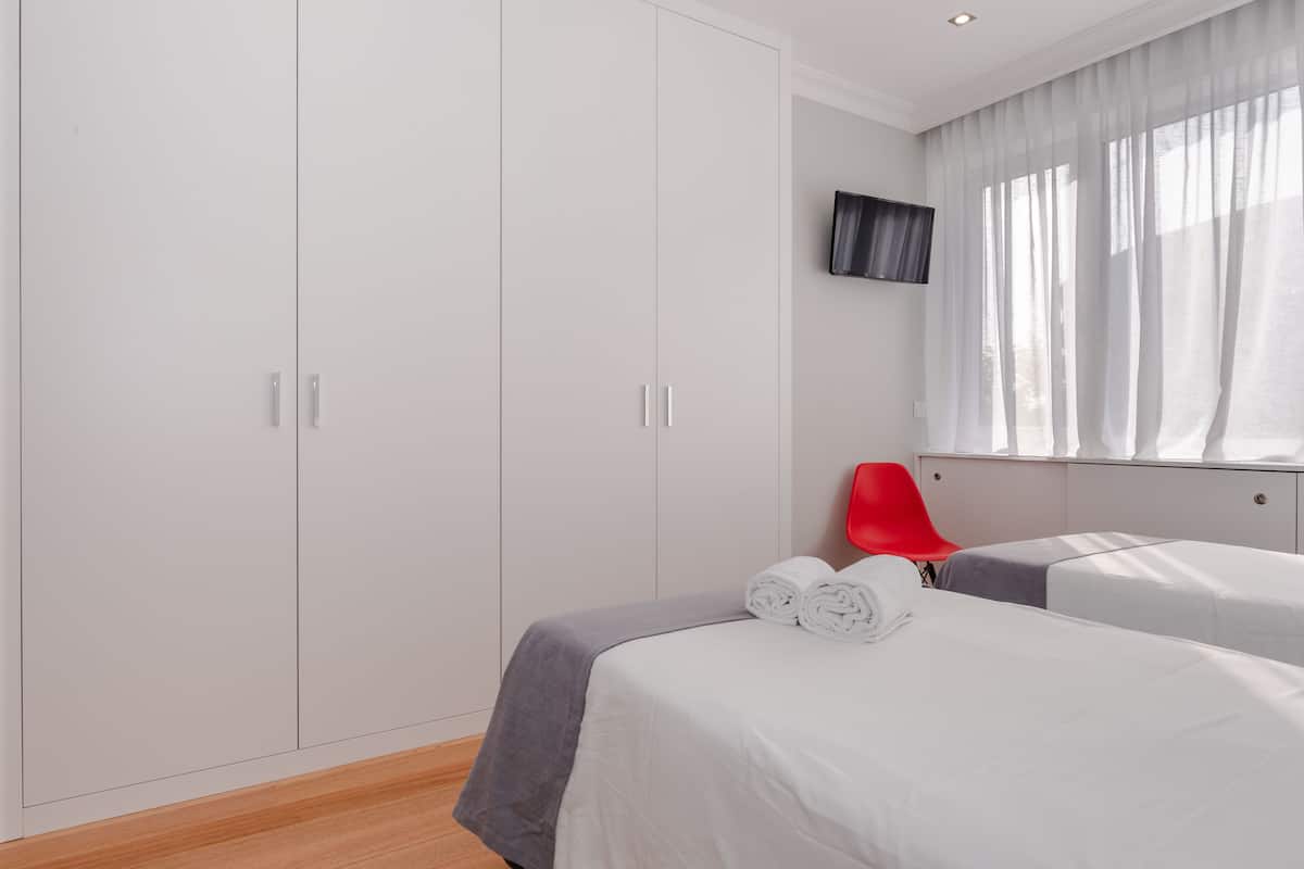 Ein Hotelzimmer mit großem Kleiderschrank, einem Bett mit grauer Bettdecke, einem roten Sessel und einem flachbildfernseher an der Wand.