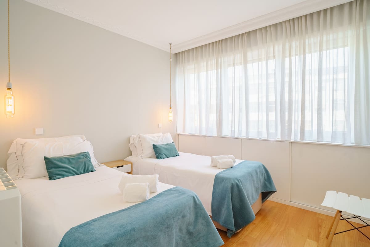 Ein Hotelzimmer mit zwei Betten, weißen Wänden und einem großen Fenster mit durchsichtigen Vorhängen.