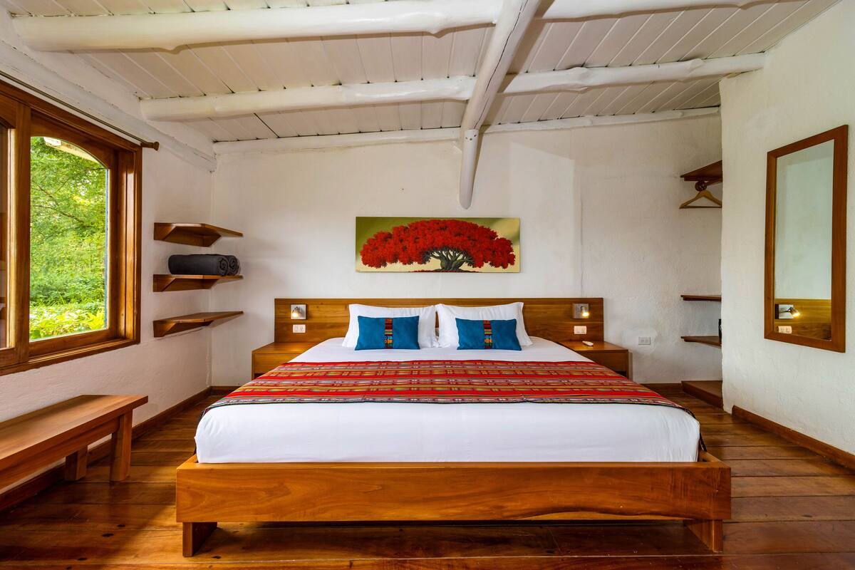 A wooden bed with white bedding and blue pillows, a painting of red flowers on the wall, wooden shelves, and a wooden bench.